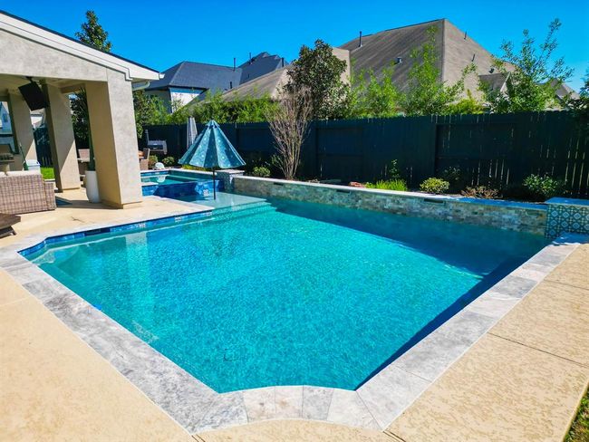 This backyard features a spacious, inviting pool with a stone-tiled border, surrounded by a landscaped area for privacy. The covered patio has comfortable seating, perfect for outdoor relaxation and entertainment. | Image 46