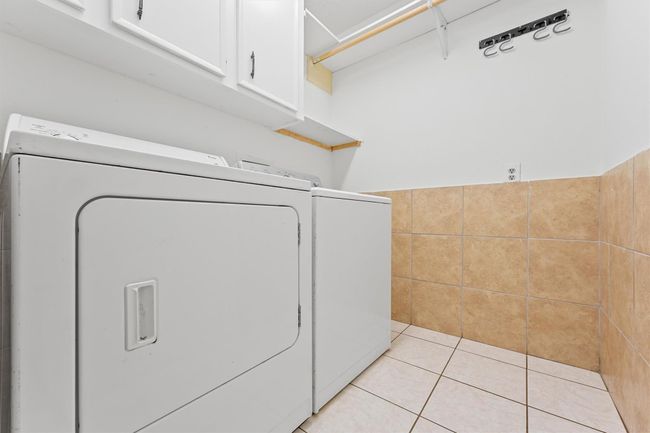 This is a laundry room featuring a washing machine and dryer. It has tiled floors and walls, overhead cabinets, and a hanging rod for convenience. | Image 13