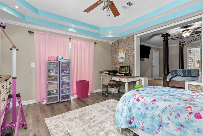 This is a spacious bedroom featuring a brick accent wall, large windows with pink curtains, and a decorative ceiling with wooden beams and blue accents. The room is currently furnished with a bed, desk, and storage units, and it opens to another room through an archway. | Image 35