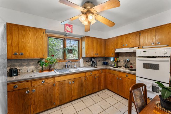 The bright kitchen is surrounded by windows and features tile flooring, tiled countertops and backsplash, and ample cabinetry—offering plenty of storage. | Image 4