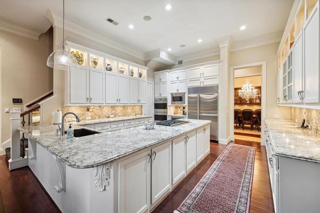 This spacious kitchen features elegant white cabinetry, granite countertops, and stainless steel appliances. A large island with a built-in cooktop and a stylish pendant light adds functionality and charm. The room is well-lit with recessed lighting and showcases a beautiful dining area view through a wide doorway. | Image 11