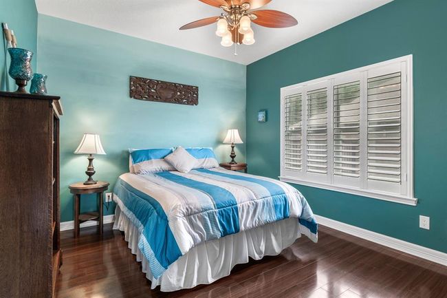 Cozy bedroom with teal walls and hardwood floors, featuring a queen-sized bed with blue and white bedding. It includes a ceiling fan, wooden furniture, and a window with white shutters, creating a bright and inviting atmosphere. | Image 22
