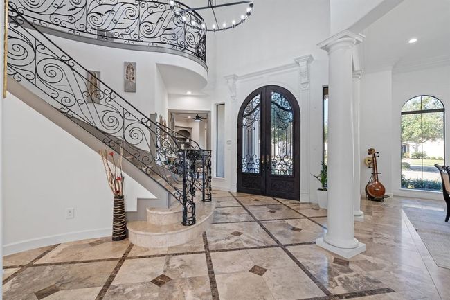 Grand entryway through the arched front door, with a striking staircase featuring a wrought iron railing and a chandelier that illuminates the space, setting an elegant tone. | Image 6