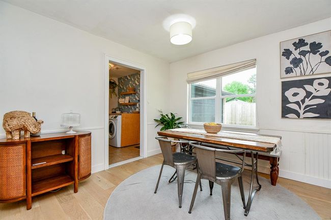 This bright dining area features a cozy wooden table with metal chairs, modern lighting, and large windows for natural light. A stylish sideboard and wall art add charm, with a view into a laundry room. | Image 22