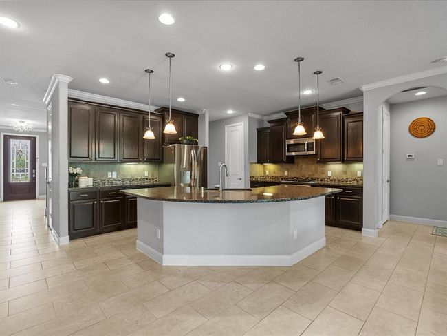 This spacious kitchen features dark wood cabinetry, a large island with pendant lighting, stainless steel appliances, and tile flooring. Adjacent is a cozy dining area, perfect for family meals. | Image 8