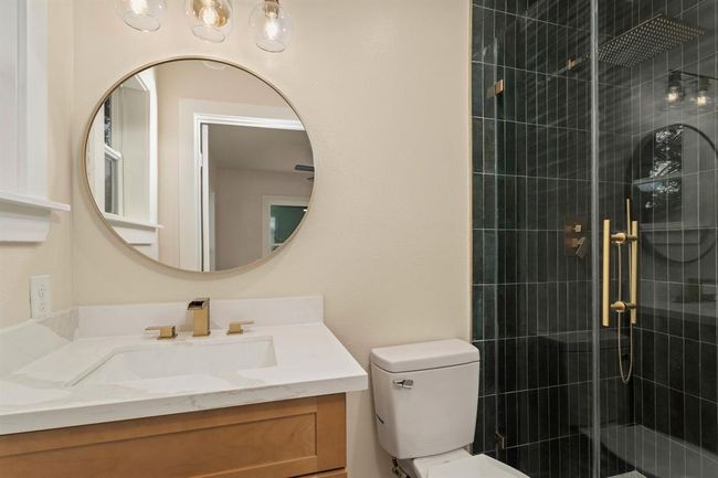 This elegant en-suite bathroom features a stylish vanity with a circular gold mirror and a coordinating light fixture overhead. A walk-in shower with a frameless glass enclosure, large rainfall showerhead, and elongated tile enhances the space, creating a refined and luxurious atmosphere. | Image 33