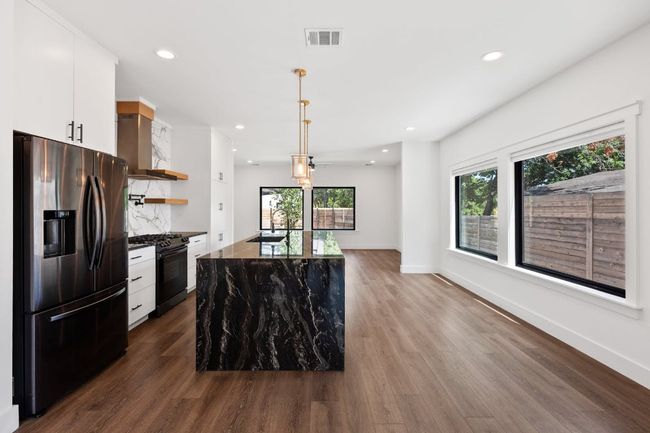 Kitchen with dark wood-style floors, black gas range oven, stainless steel fridge with ice dispenser, open shelves, and wall chimney hood | Image 7