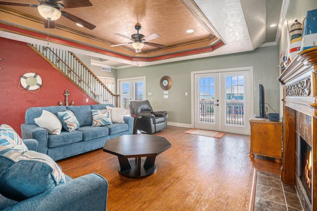 This bright and airy living room features a blend of coastal charm and comfortable living. The space is anchored by warm hardwood flooring and boasts a striking stone fireplace, adding a touch of rustic elegance. A vibrant red accent wall, adorned with nautical decor such as a life preserver and anchor, contributes to the room's character. Double doors open to reveal a deck with expansive ocean views, seamlessly connecting indoor and outdoor living spaces and filling the room with natural light. | Image 6