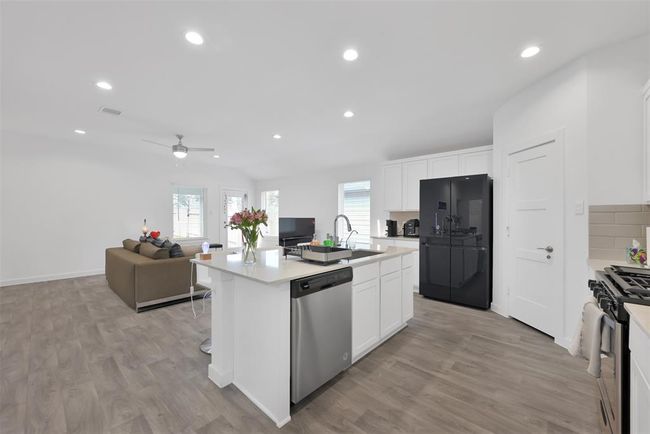 This photo showcases a modern, open-concept kitchen and living area. The kitchen features sleek white cabinets, a large island with a sink, and stainless steel appliances. The living space is bright with ample natural light, wood flooring, and a comfortable seating area, creating an inviting atmosphere for family gatherings. | Image 8