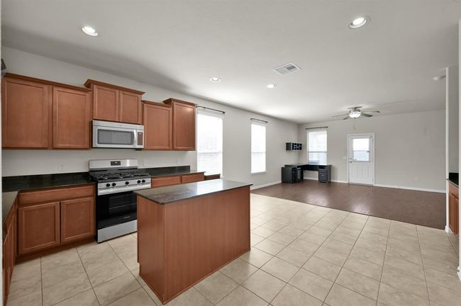 Spacious, modern kitchen with tile flooring, ample natural light, and an open layout. It features stainless steel appliances, wooden cabinetry, and a central island, leading into a well-lit living area. | Image 10