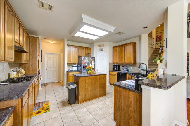 This kitchen is spacious and bright, featuring ample wooden cabinetry, a central island, and granite countertops. It has a modern gas stove, tile flooring, and a skylight for natural light. There's an open layout with easy access to adjacent rooms and stairs. | Image 12