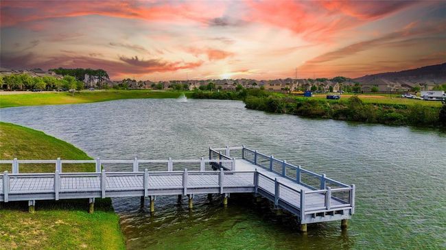 This photo showcases a serene lakeside view with a wooden dock extending over the water, set against a vibrant sunset. The surrounding area features well-maintained greenery and a nearby residential community, offering a peaceful and picturesque environment. | Image 49
