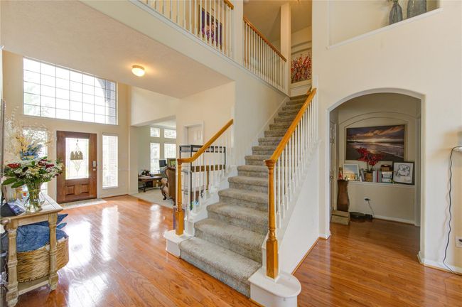 This inviting entryway features a grand staircase with carpeted steps and wooden banisters, leading to a bright, open space. The high ceilings and large windows allow natural light to flood the area, highlighting the polished hardwood floors. The front door showcases decorative glass, adding elegance to the space. | Image 16