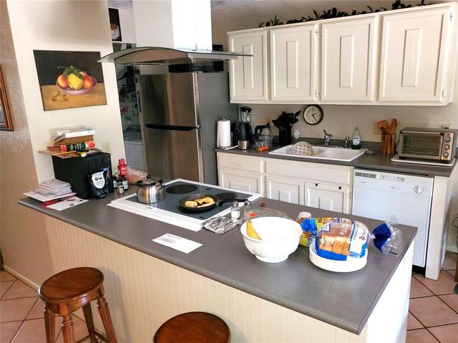 This kitchen features white cabinetry, a stainless steel refrigerator, and an island with seating. The space includes a stovetop, dishwasher, and ample counter space, providing a functional layout for cooking and entertaining. | Image 6