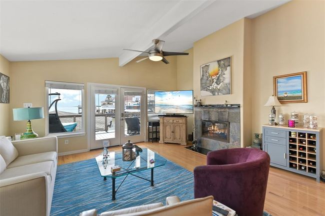 This inviting living room features large windows and glass doors leading to a deck with scenic water views. It boasts a cozy fireplace, modern ceiling fan, and comfortable seating. The space is accented with ample natural light. | Image 14