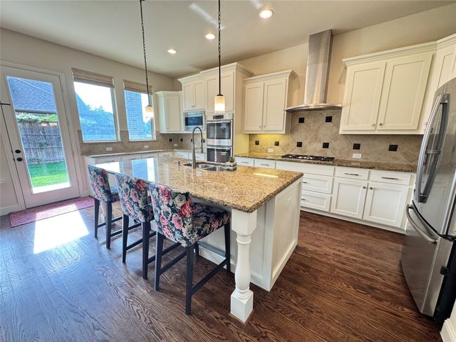 Kitchen with appliances with stainless steel finishes, dark wood finished floors, wall chimney exhaust hood, white cabinetry, and an island with sink | Image 6