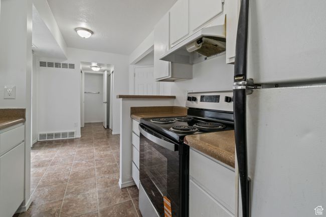 Kitchen with freestanding refrigerator, stainless steel electric range, under cabinet range hood, dark countertops, and white cabinetry | Image 5