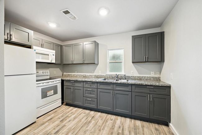 This kitchen features modern gray cabinets, granite countertops, and wood-look flooring. It includes a white refrigerator, stove, and microwave, offering a clean and contemporary design. | Image 11