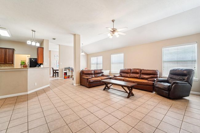 This spacious living area features tiled floors, a ceiling fan, and plenty of natural light. The open layout connects to a modern kitchen with a breakfast bar. | Image 7