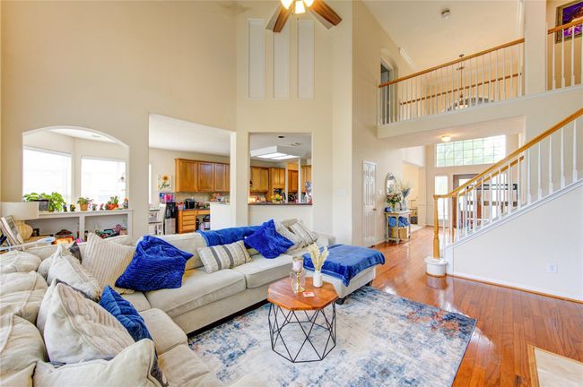 This spacious living area features high ceilings and an open floor plan, connecting seamlessly to the kitchen. It boasts abundant natural light, stylish wood flooring, and a cozy sectional sofa, perfect for family gatherings. A staircase leads to the upper level, adding to the home's elegant design. | Image 18