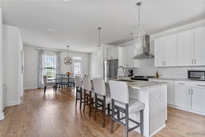 Kitchen with wall chimney range hood, light wood-type flooring, a center island with sink, and stainless steel appliances | Image 3