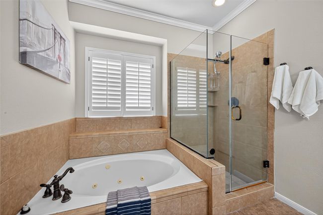 This bathroom features a luxurious jetted tub and a separate glass-enclosed shower. It has elegant beige tile work, a large window with plantation shutters, and tasteful decor, creating a serene and inviting space. | Image 32