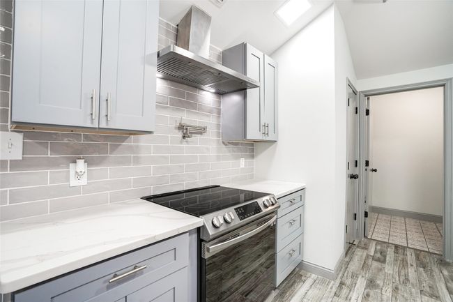 Kitchen with stainless steel electric stove, wall chimney exhaust hood, light stone counters, light wood finished floors, and decorative backsplash | Image 6