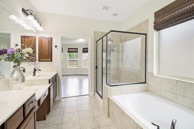 The spacious bathroom with a modern design, featuring a large glass-enclosed shower, a separate bathtub, dual sinks with ample counter space, and elegant cherry wood cabinetry. Natural light filters through a window with a woven shade, enhancing the bright, airy atmosphere. | Image 15