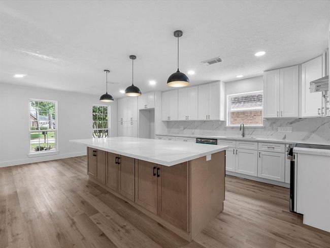 This photo showcases a spacious, modern kitchen with a large central island featuring a sleek white countertop. The kitchen is equipped with white cabinetry and stainless steel appliances, complemented by wood flooring and pendant lighting. Large windows provide ample natural light. | Image 16