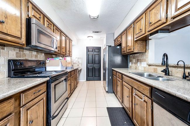 This kitchen features a galley layout with wooden cabinetry, stainless steel appliances, and a double sink. The countertops are granite, and the backsplash is tiled. It has a bright, open feel with overhead lighting and light-colored floor tiles. | Image 11