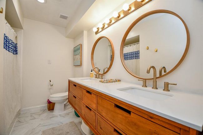 This bathroom features a modern design with dual round mirrors, a wooden vanity with double sinks, and brushed gold fixtures. The space is brightened by ample lighting and accented with a patterned shower curtain, creating a stylish and functional environment. | Image 5