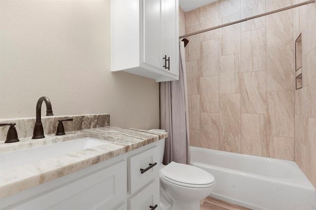 This bathroom features a sleek design with a marble countertop, dark fixtures, white cabinetry, and a tiled shower-tub combo, offering a modern and clean look. | Image 10
