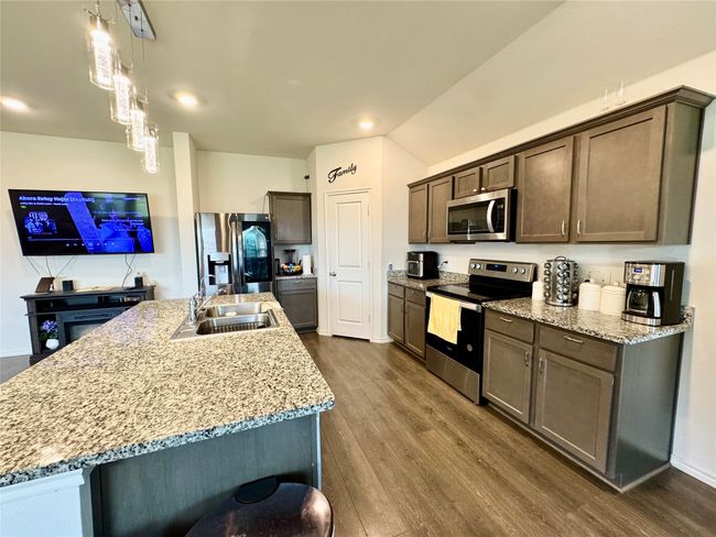 Kitchen with stainless steel appliances, dark wood-style floors, dark brown cabinetry, an island with sink, and light stone countertops | Image 5