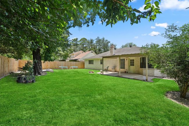 This photo shows a spacious backyard with lush green grass, a large tree, and a wooden fence for privacy. There's a cozy covered patio attached to the house, ideal for outdoor relaxation or entertaining. | Image 26