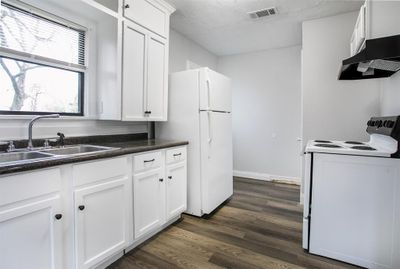 This is a bright, modern kitchen featuring white cabinetry, dark countertops, and updated appliances. There's a window above the sink for natural light and wood-look flooring throughout. | Image 2