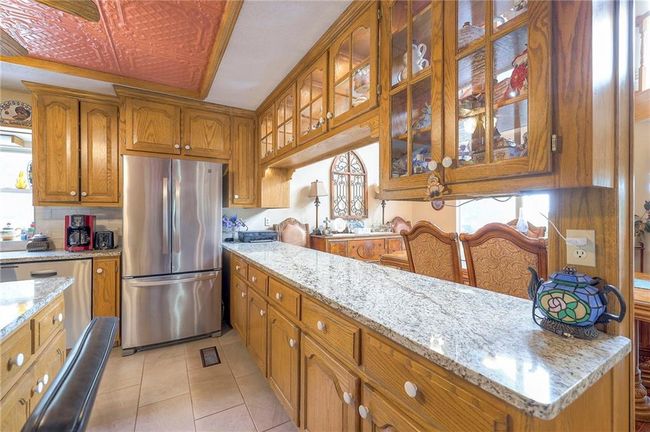 Kitchen featuring appliances with stainless steel finishes, brown cabinets, light tile patterned floors, glass insert cabinets, and light stone counters | Image 14
