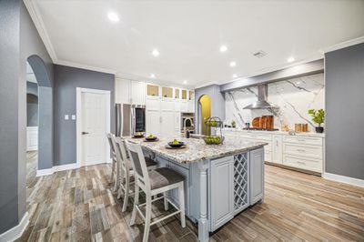 This is a spacious, modern kitchen featuring white cabinetry, granite countertops, and a large center island with seating. It has stainless steel appliances, wood-look tile flooring, and a stylish marble backsplash. The room is well-lit with recessed lighting and offers a neutral color scheme with gray accents. | Image 3