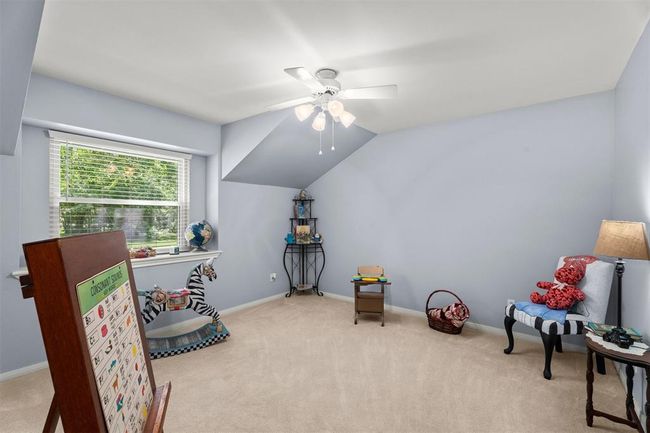 This room features a cozy, carpeted space with soft gray walls and a sloped ceiling. A large window lets in natural light, and a ceiling fan provides comfort. Ideal for a child's playroom or a creative space, it includes playful decor elements and ample floor space. | Image 17