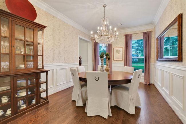 To the left of the entry, an elegant formal dining room awaits. This space exudes classic charm with double crown moulding, wainscoting with chair rail, and timeless wallpaper accents. An exquisite chandelier hangs as a focal point, complemented by two large front-facing windows that invite natural light and enhance the room’s welcoming ambiance. | Image 7