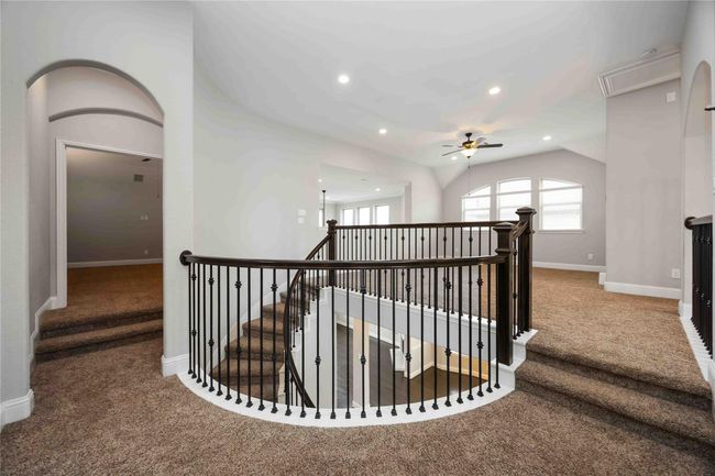 Expansive upstairs landing highlighted by elegant wrought iron railings, soft plush carpeting, and recessed lighting. The open layout includes a ceiling fan and large windows that fill the space with abundant natural light. | Image 30