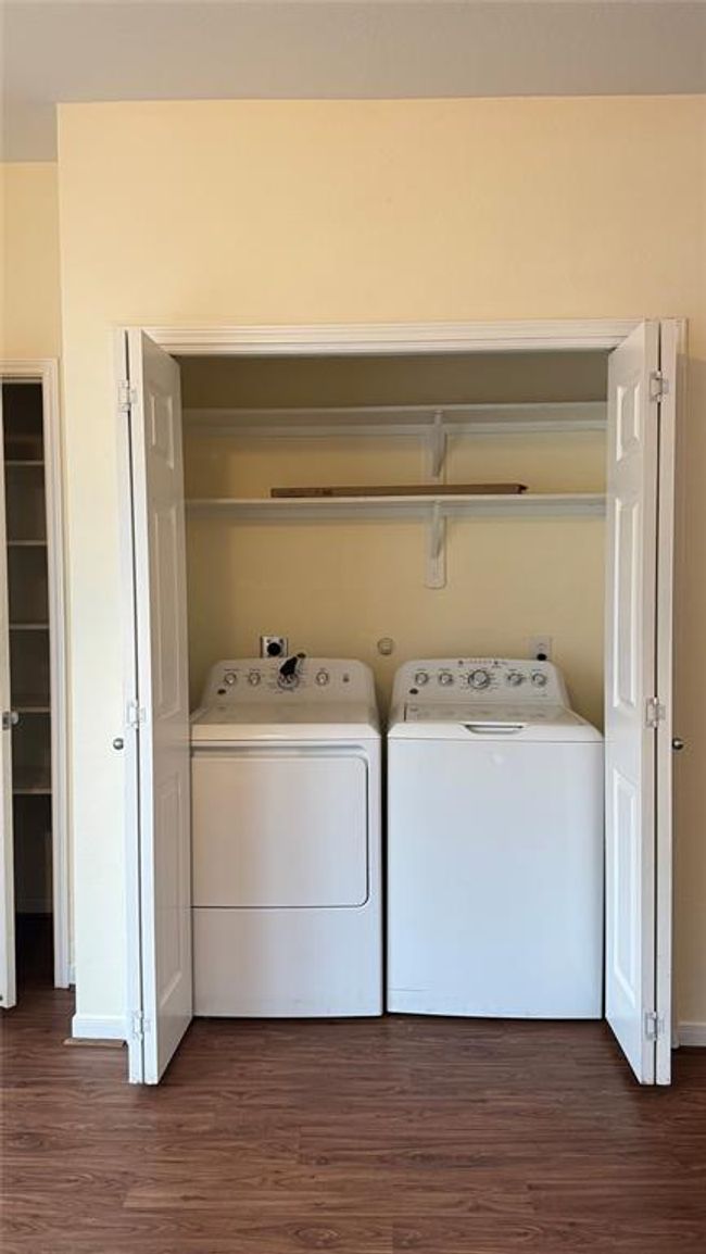This photo shows a compact laundry area with a washer and dryer neatly tucked into a closet space. It features bi-fold doors for easy access and a shelf above for storage. The area has a clean, organized look with light-colored walls and wood flooring. | Image 7