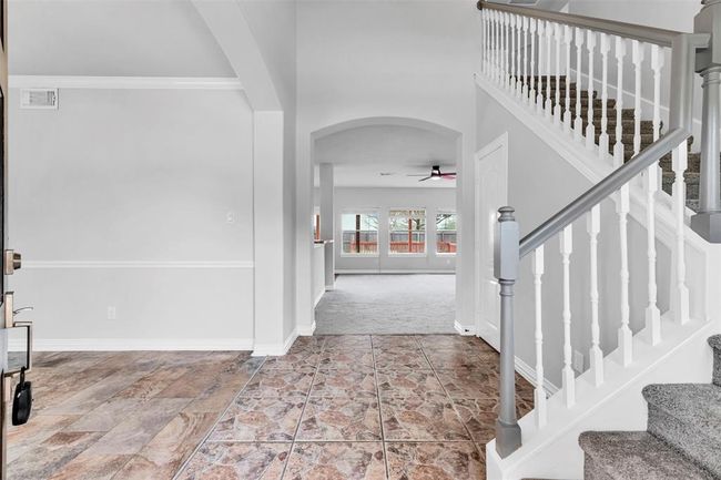 This photo showcases a bright entryway with tiled flooring leading to a carpeted living area. The space features a white staircase with elegant railing, an arched doorway, and large windows providing ample natural light. | Image 5