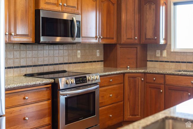 Kitchen with appliances with stainless steel finishes, brown cabinetry, decorative backsplash, and light stone countertops | Image 11