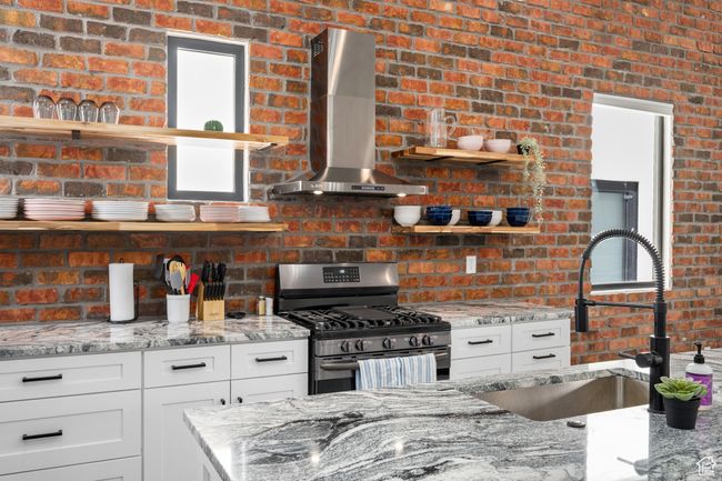 Kitchen with brick wall, wall chimney exhaust hood, sink, light stone counters, and stainless steel range with gas stovetop | Image 33