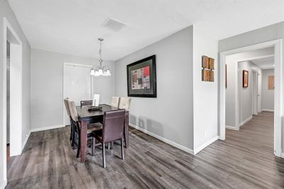 This is a modern dining area with wood-look flooring, light gray walls, and a classic chandelier. The space features a dark wood dining table with chairs, and there is easy access to adjacent rooms, offering a sense of flow and openness. | Image 3