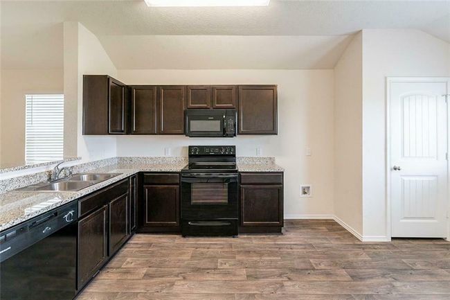 Kitchen with black appliances, vaulted ceiling, light wood finished floors, dark brown cabinets, and light stone countertops | Image 5