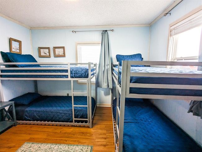 This bedroom features two sets of bunk beds with blue bedding, ideal for maximizing space. The room is well-lit with natural light from two windows and has light blue walls, creating a bright and inviting atmosphere. | Image 16