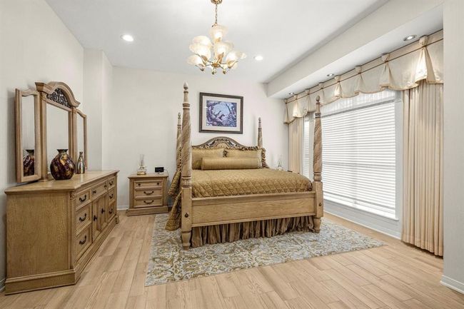 This is a spacious and elegant bedroom featuring a large four-poster bed, matching wooden dresser and nightstand, and a full-length mirror. The room has light-colored walls and hardwood flooring, complemented by an ornate area rug. A window with stylish drapes allows for natural light, and a classic chandelier adds a touch of sophistication. | Image 24