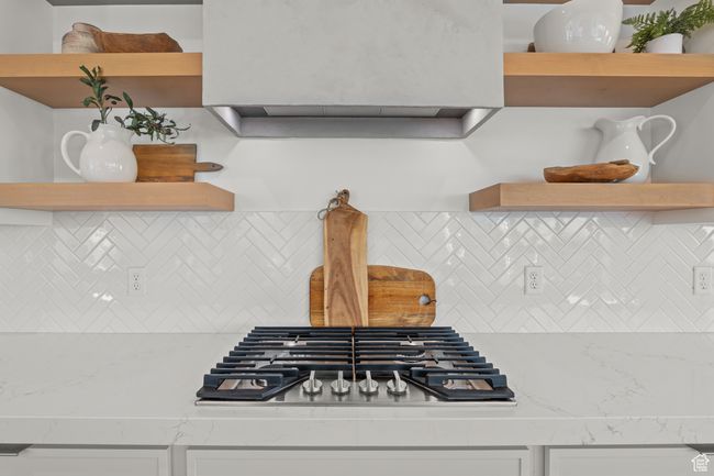 Kitchen view of backsplash, stainless steel gas cooktop, under cabinet range hood, light stone countertops, and open shelves | Image 19