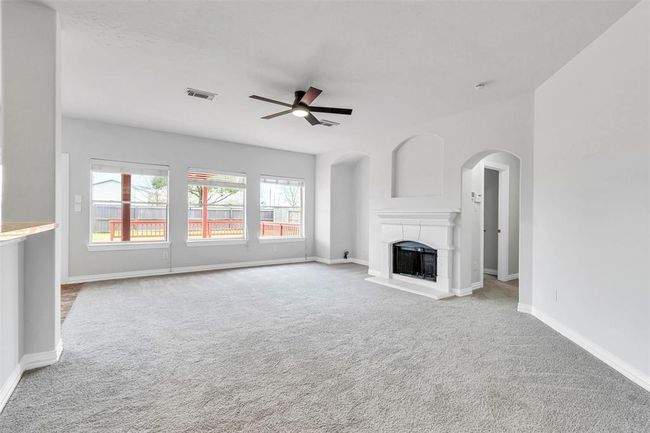 This spacious living room features soft gray carpeting, a modern ceiling fan, and ample natural light from large windows. It includes a stylish white fireplace and has an open, airy feel with neutral tones. | Image 8